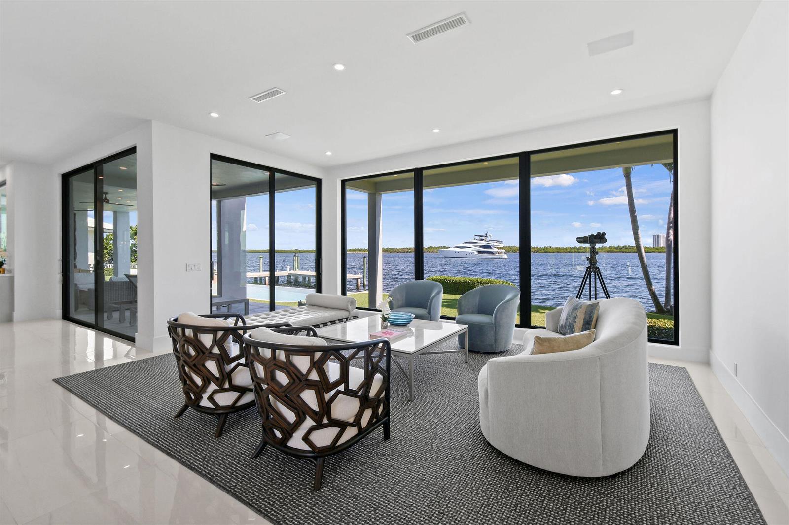 A modern living room features floor-to-ceiling windows overlooking a waterfront view with a yacht — 101 Bowsprit Drive, North Palm Beach FL 33408, NORTH PALM BEACH, FL