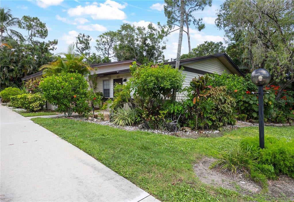 A single-story house with light siding is surrounded by lush landscaping and a concrete walkway — 1001 PINE ISLE LANE UNIT 1001, NAPLES, FL