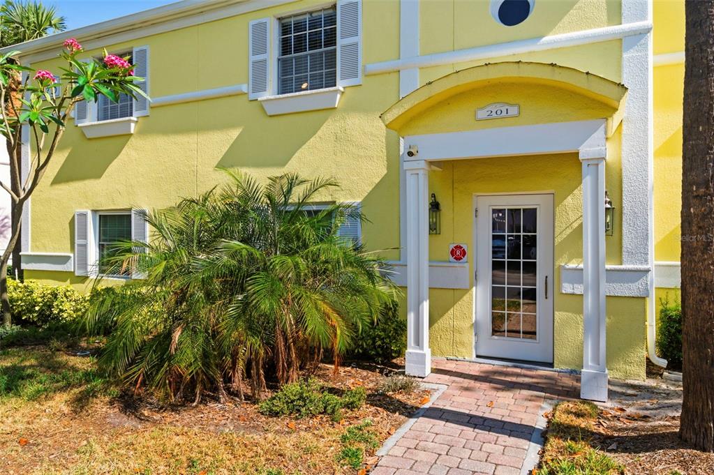 A yellow two-story building exterior features a brick walkway leading to a white front door — 201 POMPANO DRIVE SE UNIT C furnished, ST PETERSBURG, FL