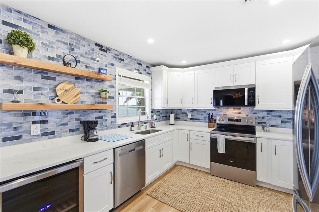 A kitchen features white cabinets, stainless steel appliances, a blue tiled backsplash, and wood shelving — 201 POMPANO DRIVE SE UNIT C furnished, ST PETERSBURG, FL