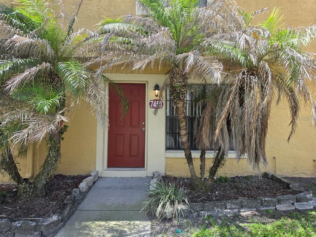 A red front door is flanked by two palm trees in front of a yellow building — 7450 WYNNEWOOD SQUARE UNIT 189, WINTER PARK, FL