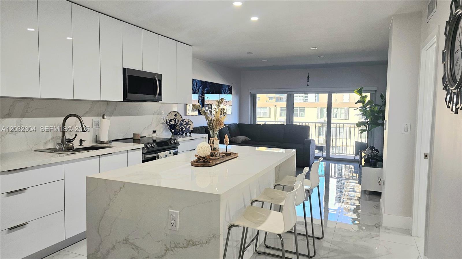 A modern kitchen features white cabinetry, a marble-topped island with bar stools, and living space — 1950 S Ocean Dr # 3D, Hallandale Beach FL 33009, HALLANDALE BEACH, FL