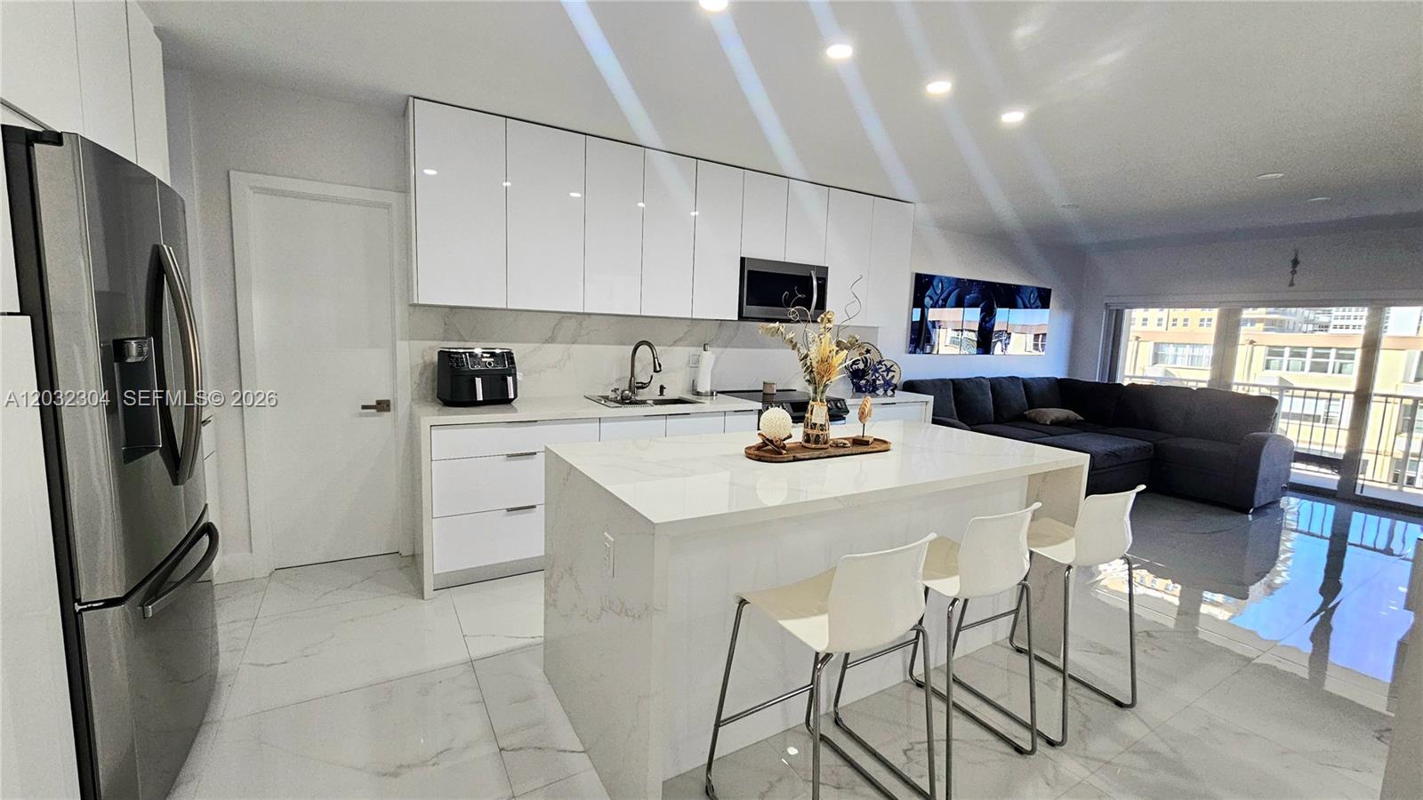 A modern kitchen features white cabinetry, a marble-topped island, and an adjacent living area — 1950 S Ocean Dr # 3D, Hallandale Beach FL 33009, HALLANDALE BEACH, FL