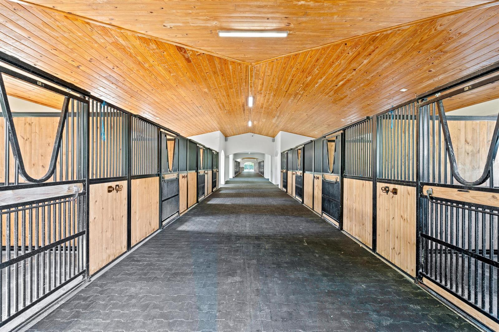 A horse stable aisle with wooden ceilings, black metal stall doors, and paved flooring — 5656 South Shore Boulevard, Wellington FL 33449, WELLINGTON, FL