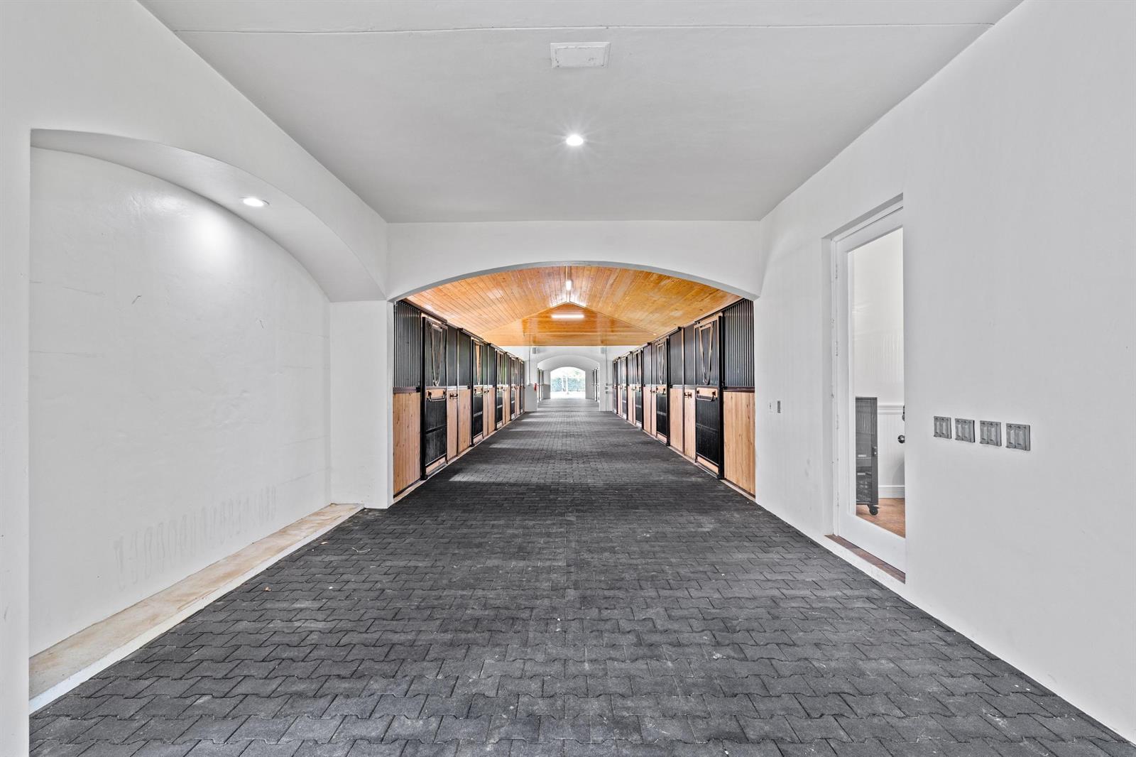 A long, covered stable aisle features dark brick flooring, wooden stall doors, and arched ceilings — 5656 South Shore Boulevard, Wellington FL 33449, WELLINGTON, FL