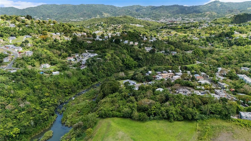 An aerial view shows a river flowing through a lush, hilly landscape with scattered houses — TRAZO BARRIO RINCON, CIDRA, PR