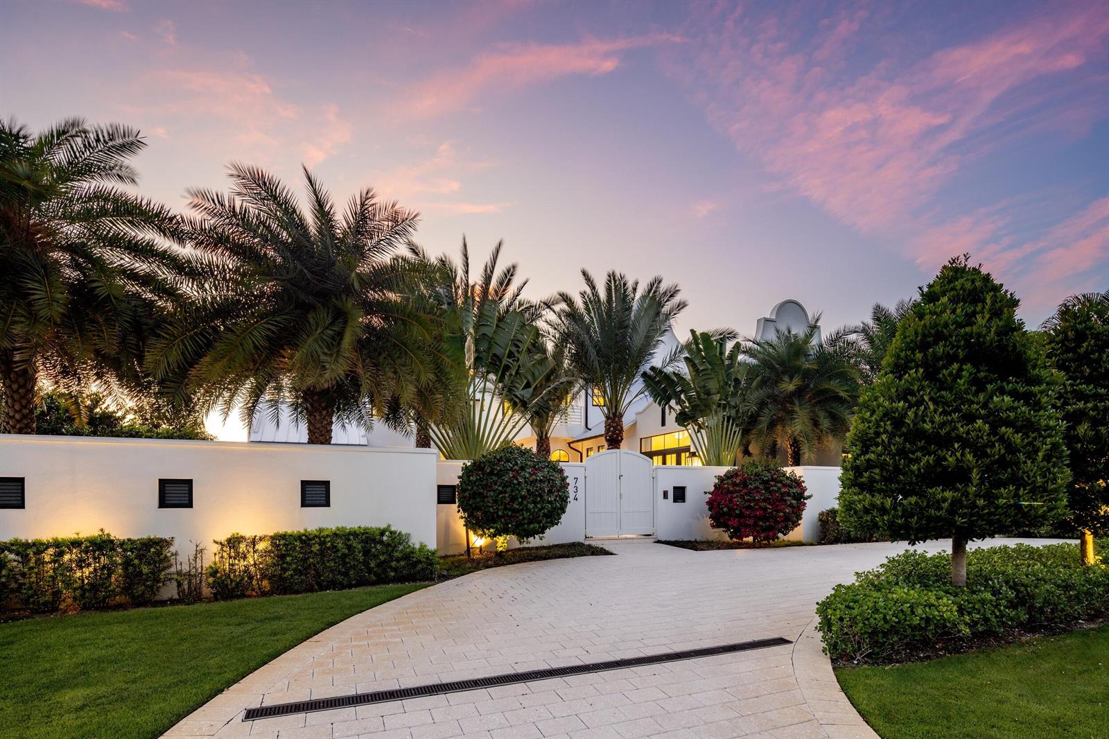 A white walled exterior with a paved driveway, palm trees, and a gated entrance at dusk — 734 S Ocean Boulevard, Delray Beach FL 33483, DELRAY BEACH, FL