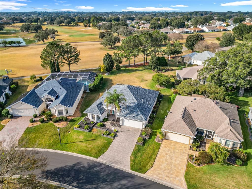 An aerial view shows three suburban houses with driveways and lawns bordering a golf course — 16816 SE 85TH SAPELO COURT, THE VILLAGES, FL