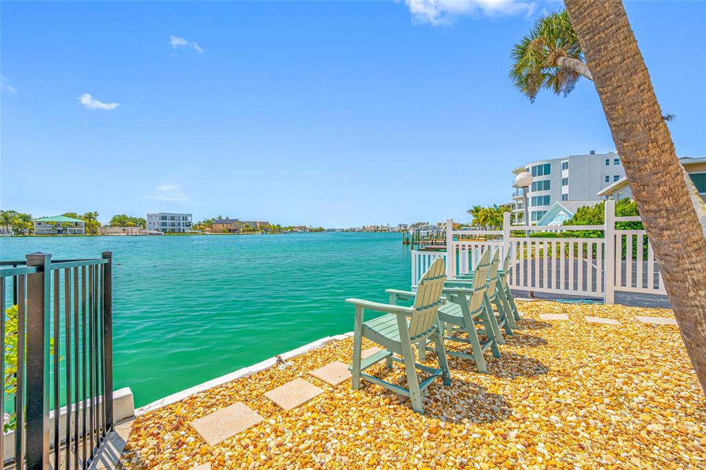 A waterfront patio features light blue Adirondack chairs on a gravel surface with a fence — 285 107TH AVENUE UNIT 306, TREASURE ISLAND, FL