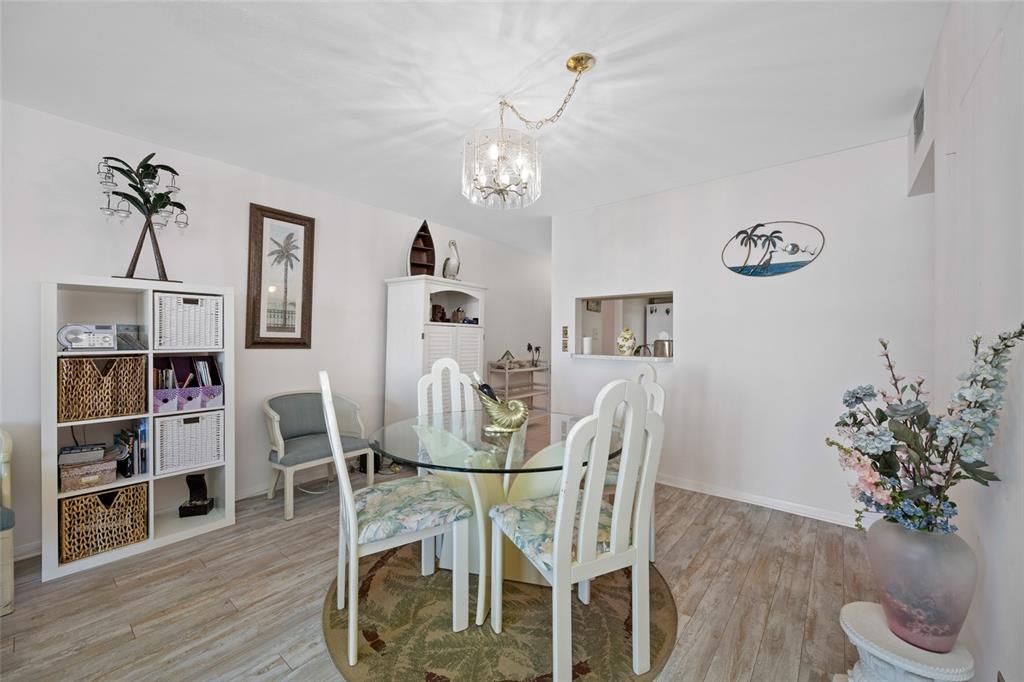 A dining room features a glass-topped table, white chairs, a bookshelf, and light wood flooring — 285 107TH AVENUE UNIT 306, TREASURE ISLAND, FL