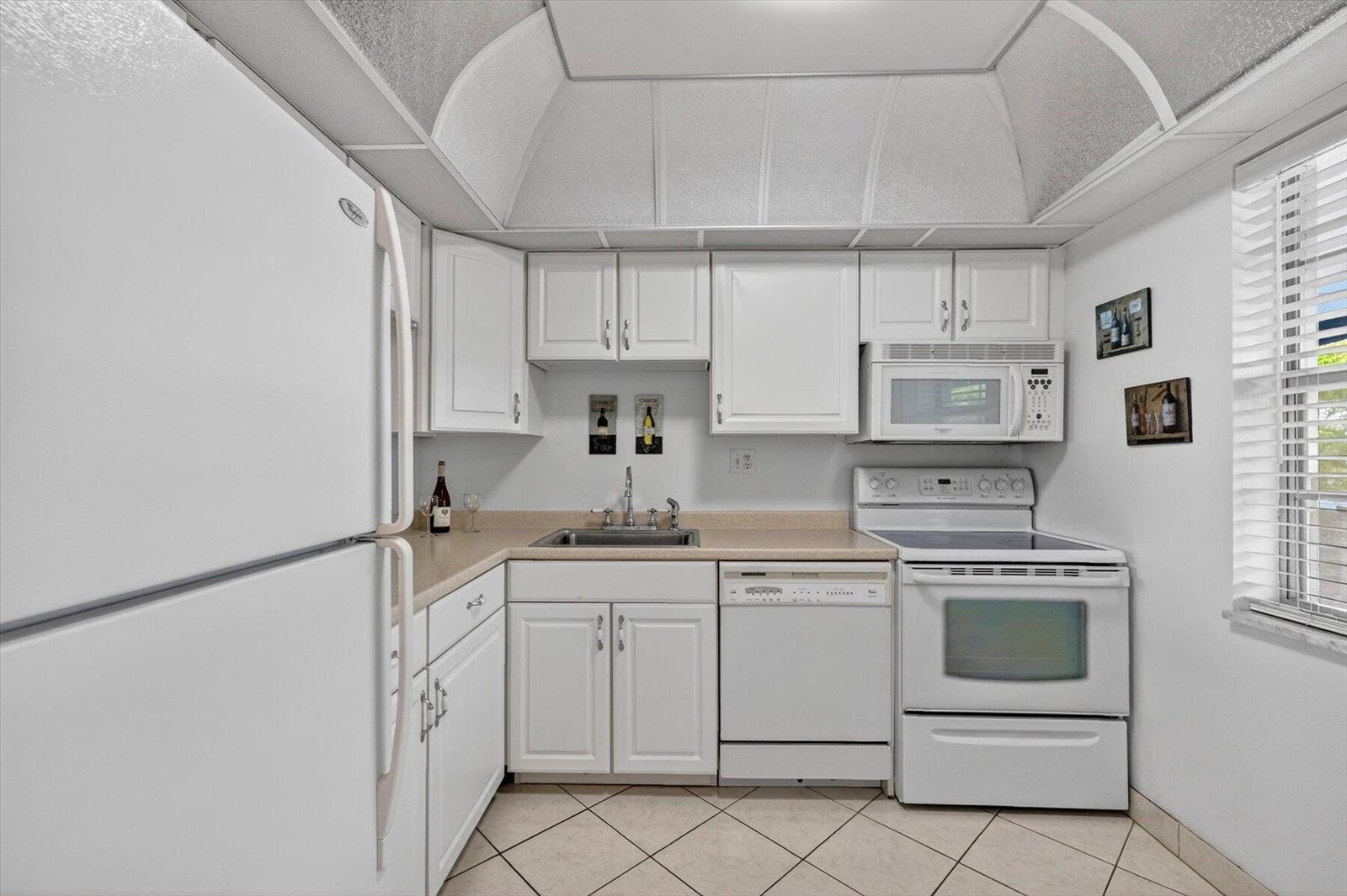 A small kitchen features white cabinets, white appliances, a beige countertop, and a vaulted ceiling — 7787 Golf Circle Drive # k, Margate FL 33063, MARGATE, FL