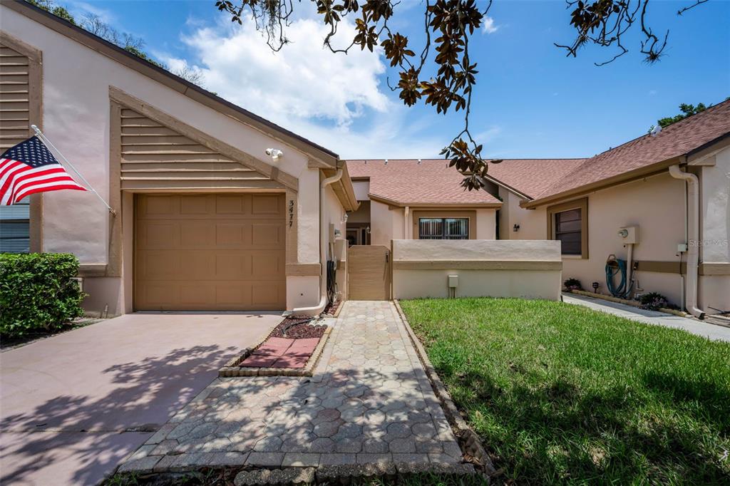 A tan house exterior features a garage, a paved walkway, and an American flag — 3477 HILLMOOR DRIVE, PALM HARBOR, FL