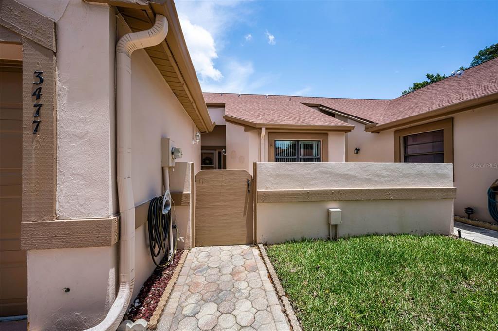 A paved walkway leads to a wooden gate and the entrance of a stucco house — 3477 HILLMOOR DRIVE, PALM HARBOR, FL