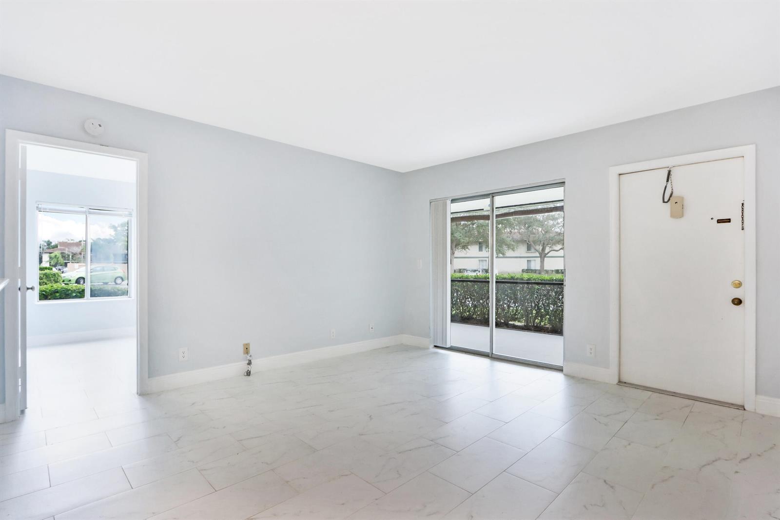 An empty living room features light-colored tile floors, white walls, a sliding glass door, and an entryway — 6313 Chasewood Drive # B, Jupiter FL 33458, JUPITER, FL