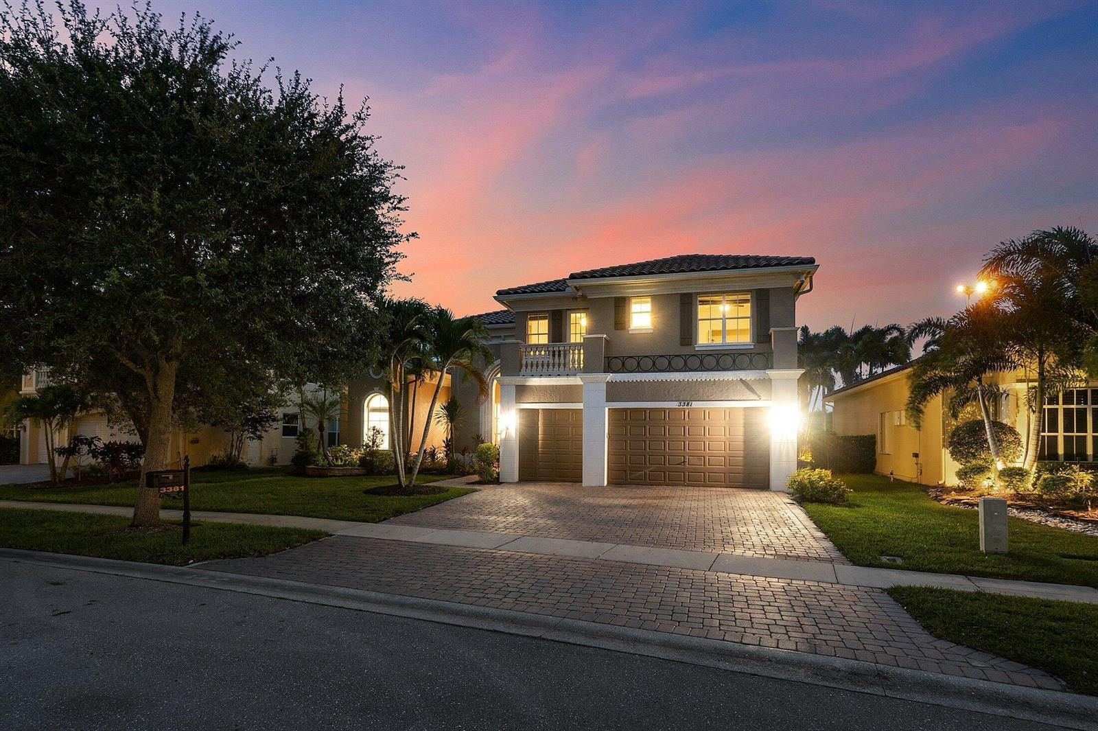 A two-story house with a paved driveway, a three-car garage, and palm trees at sunset — 3381 Lago De Talavera, Lake Worth FL 33467, LAKE WORTH, FL