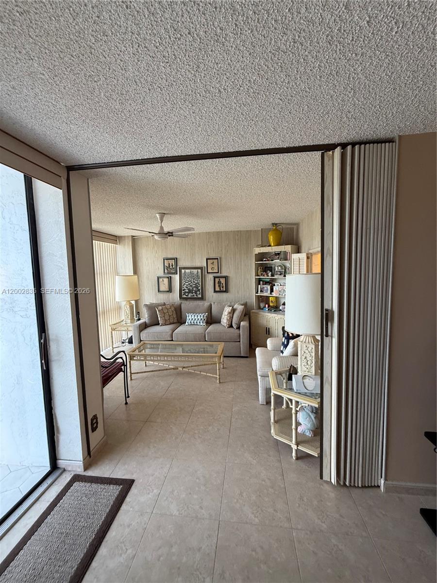A living room features a beige sofa, tiled floors, a ceiling fan, and accordion doors — 1000 Parkview Dr # 407, Hallandale Beach FL 33009, HALLANDALE BEACH, FL