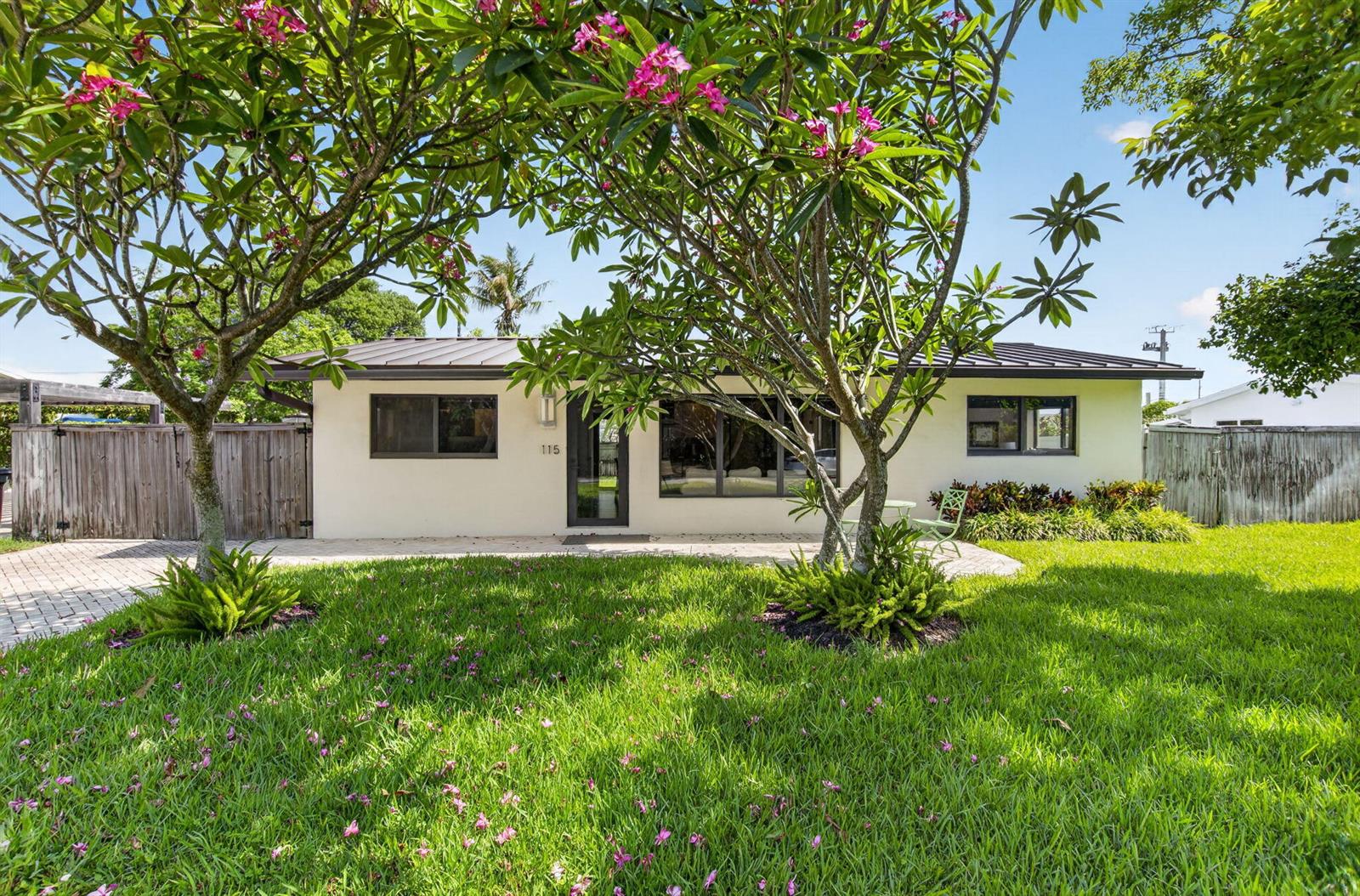 A single-story house with a light-colored exterior, dark window frames, and two flowering plumeria trees — 115 NW 7th Street, Boca Raton FL 33432, BOCA RATON, FL