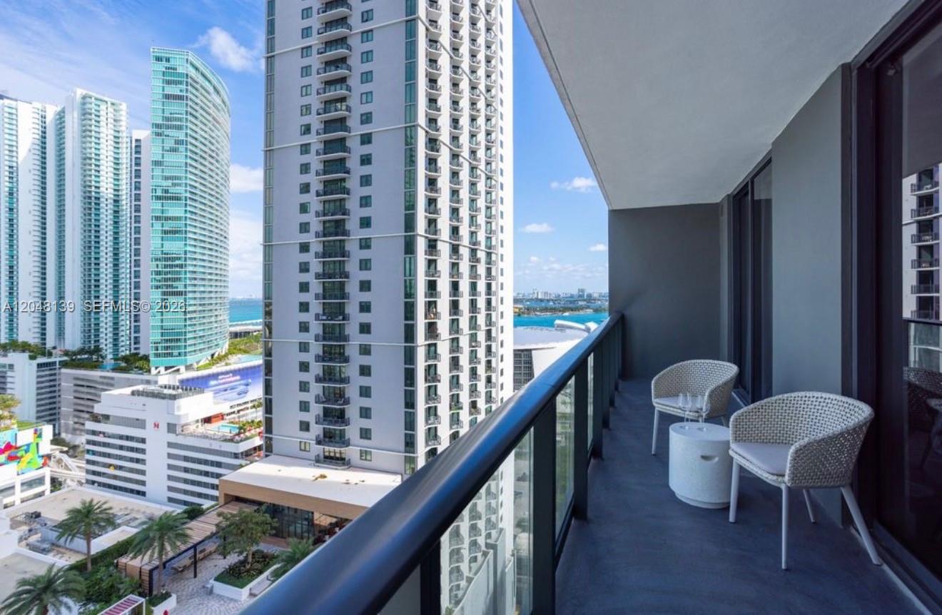 A high-rise balcony features two wicker chairs, a small table, and views of city skyscrapers — 159 NE 6th St # 2002, Miami FL 33132, MIAMI, FL