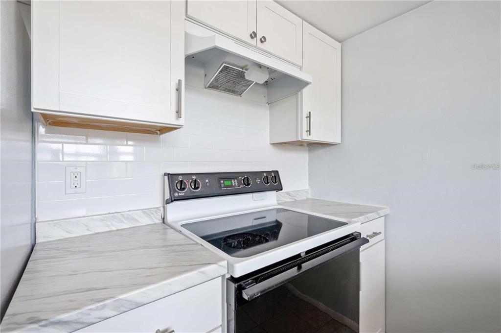 A kitchen features white cabinets, a glass-top stove, and marble-patterned countertops with a subway tile backsplash — 1433 S BELCHER ROAD UNIT G16, CLEARWATER, FL
