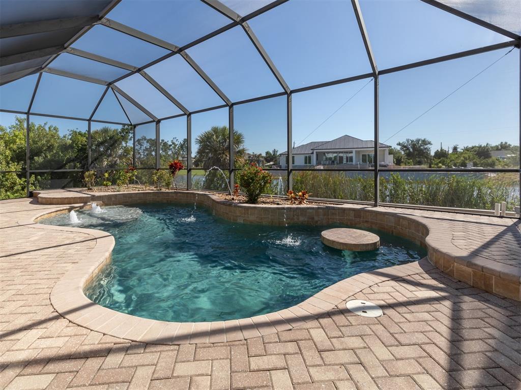 A screened-in swimming pool with fountains and a stone patio overlooks a lake and house — 14462 FORT WORTH CIRCLE, PORT CHARLOTTE, FL
