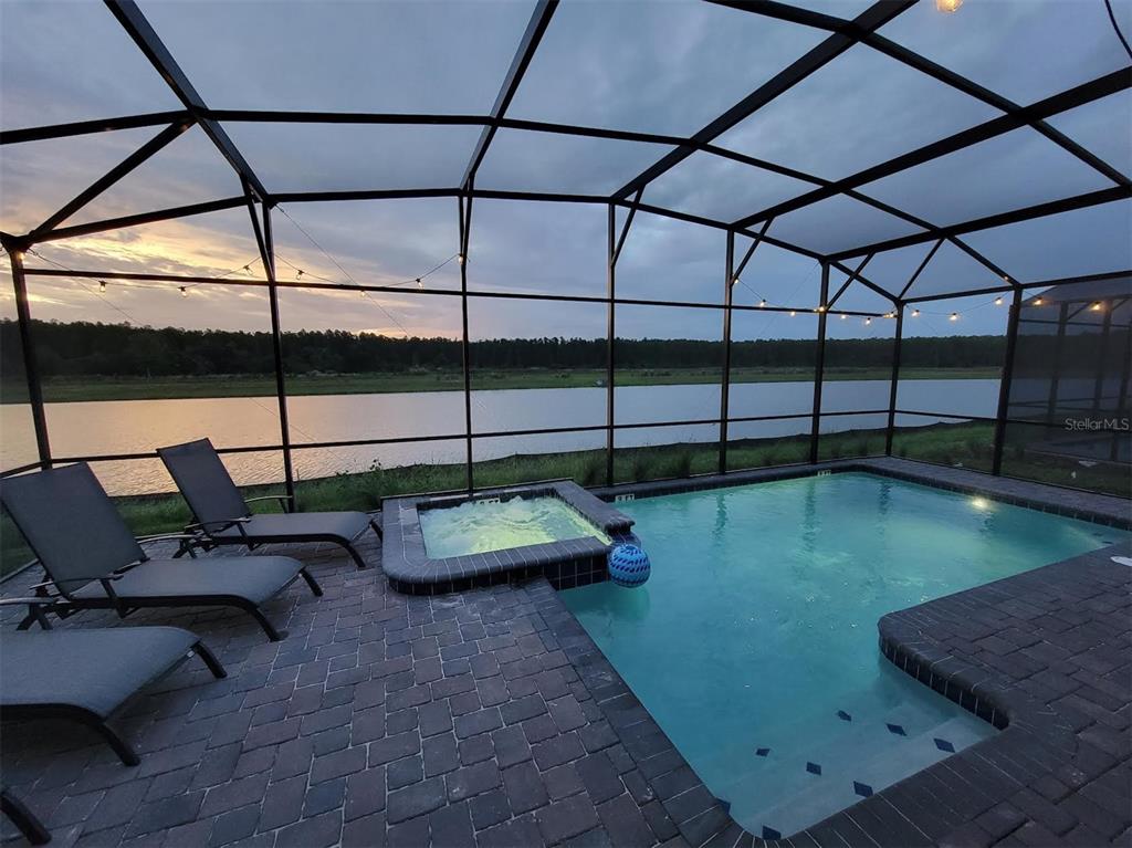 A screened-in pool and spa overlook a lake at sunset with lounge chairs on the deck — 4173 PARAGRAPH DRIVE, KISSIMMEE, FL