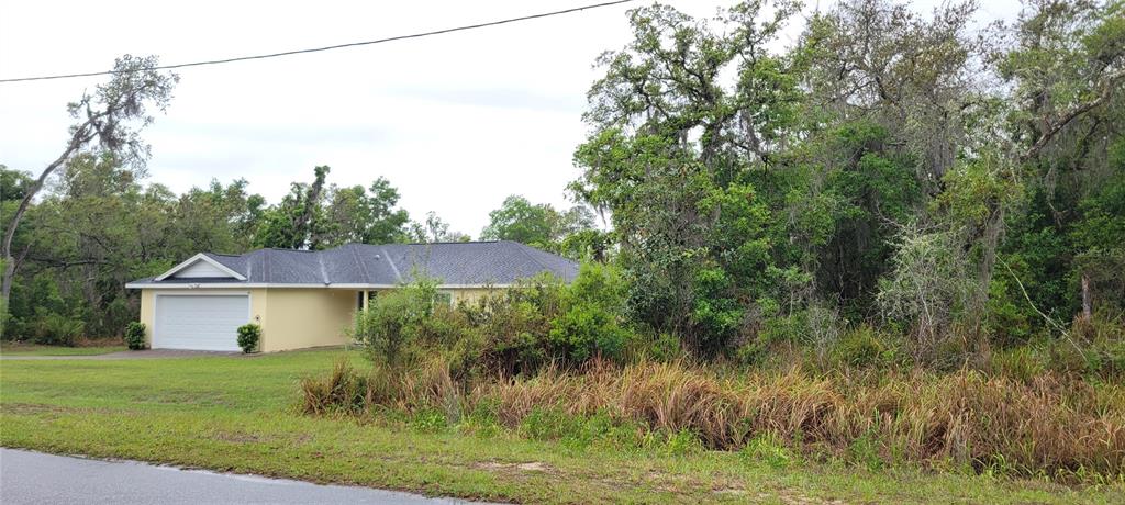 A yellow single-story house with a two-car garage sits behind a grassy, wooded lot — SPRUCE ROAD, OCALA, FL