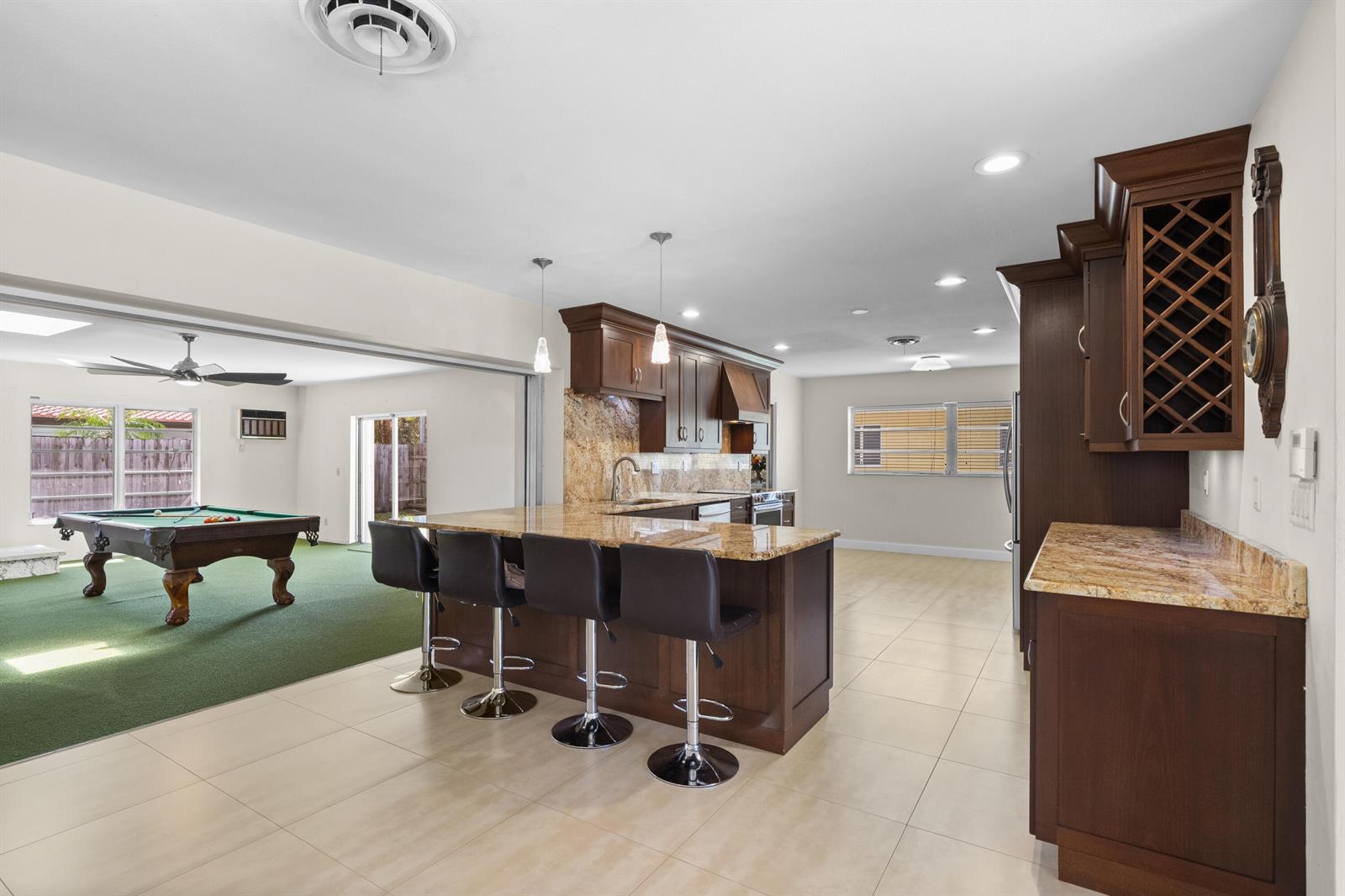 A kitchen with dark wood cabinets and granite countertops overlooks a room with a pool table — 1500 SE 24th Avenue, Pompano Beach FL 33062, POMPANO BEACH, FL