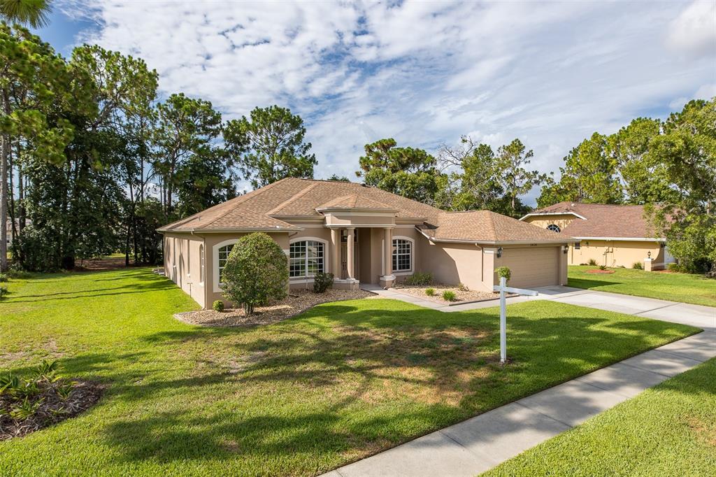 A single-story tan stucco house with a brown shingled roof and a front yard — 5436 CHAMPIONSHIP CUP LANE, SPRING HILL, FL