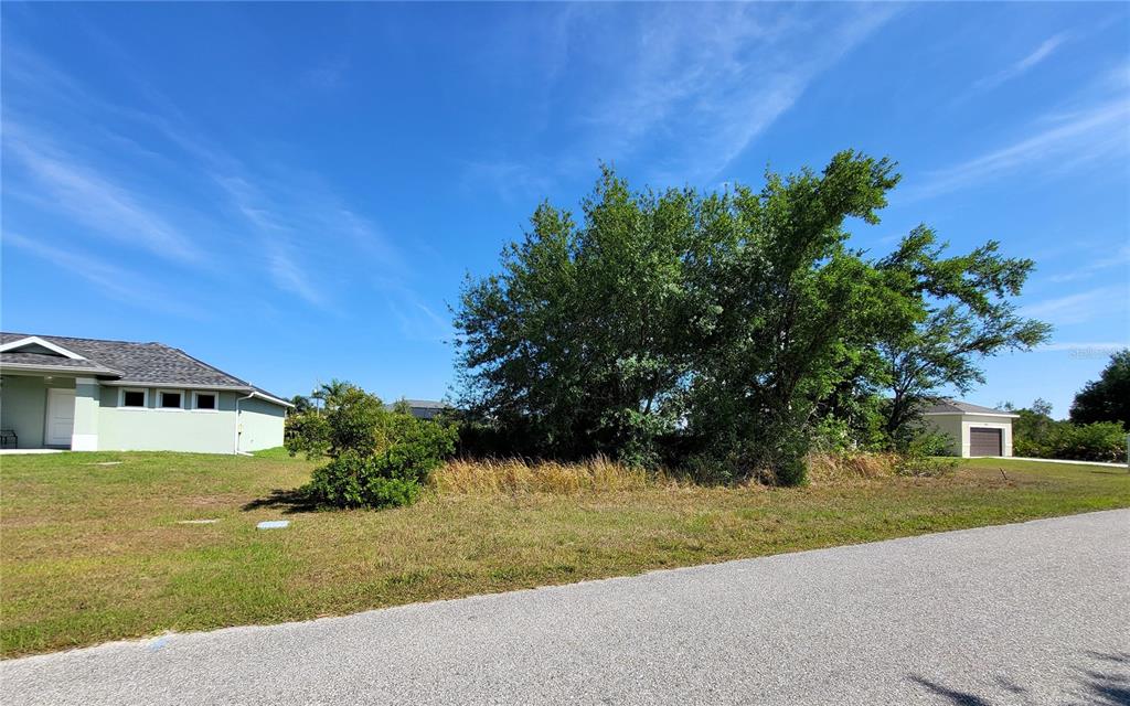 A vacant, grassy lot with trees sits between two suburban houses along a paved road — 9164 RAVEL STREET, PORT CHARLOTTE, FL