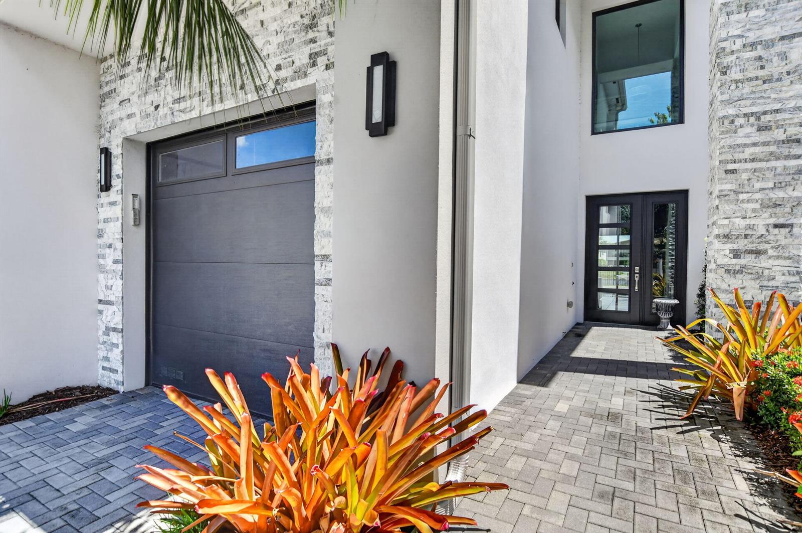 A modern home exterior features a dark garage door, stone accents, and a paved walkway — 17302 Rainstream Road, Boca Raton FL 33496, BOCA RATON, FL