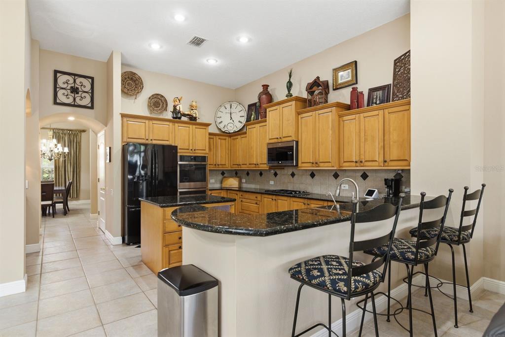 A kitchen features light wood cabinets, black granite countertops, stainless steel appliances, and bar seating — 5442 MILLBROOK WAY, PALM HARBOR, FL