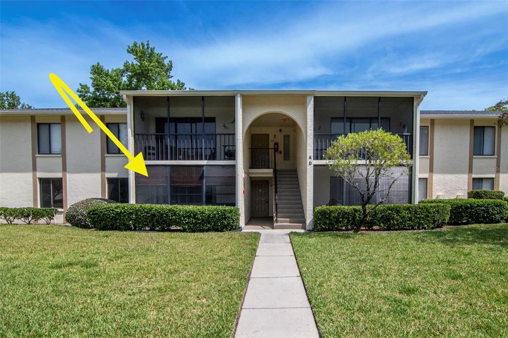 A two-story apartment building exterior features a ground-floor screened patio with a yellow arrow — 3211 LAKE PINE WAY E UNIT A1, TARPON SPRINGS, FL
