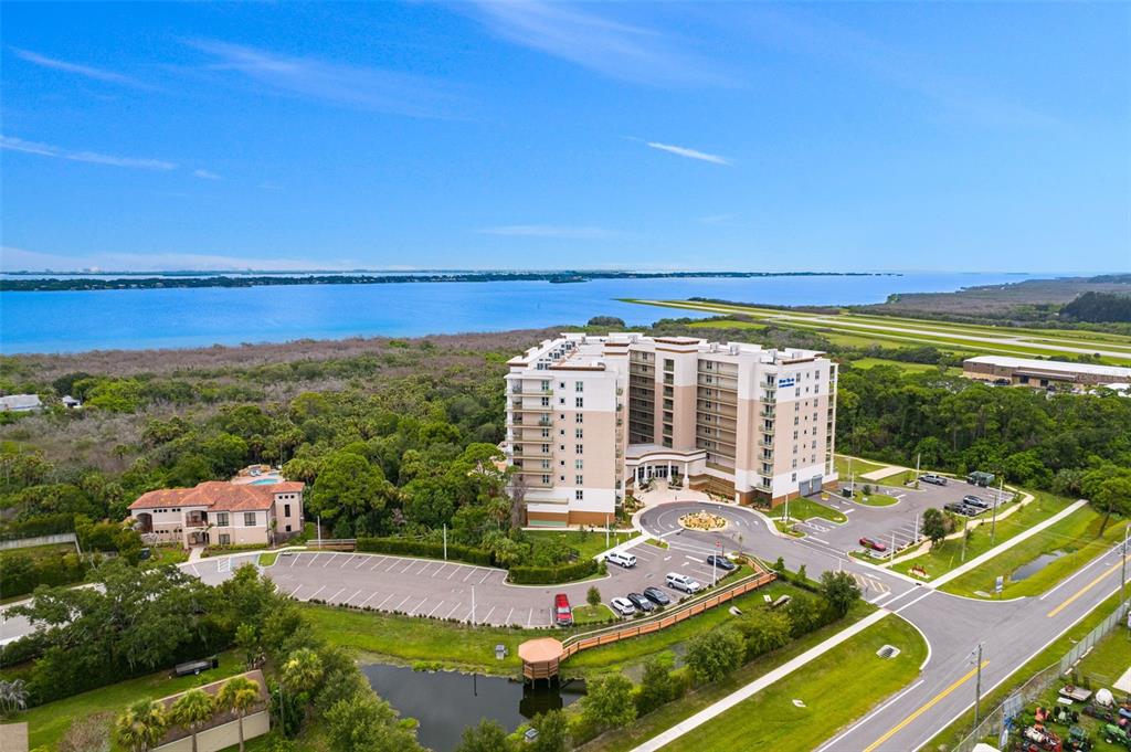 An aerial view shows a multi-story apartment building surrounded by trees, a parking lot, and water — 735 W PILOT LANE UNIT 506, MERRITT ISLAND, FL