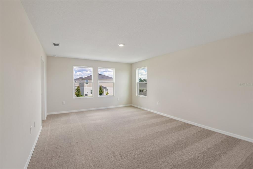 An empty bedroom features beige carpet, light-colored walls, and three windows overlooking a neighborhood — 4192 RIVER BEACON BOULEVARD, DAVENPORT, FL