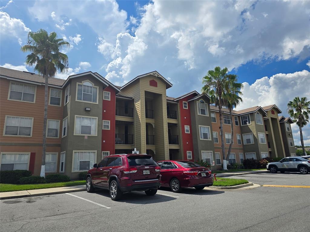 A multi-story apartment building with tan and red siding stands behind a parking lot — 2206 ANTIGUA PLACE, KISSIMMEE, FL