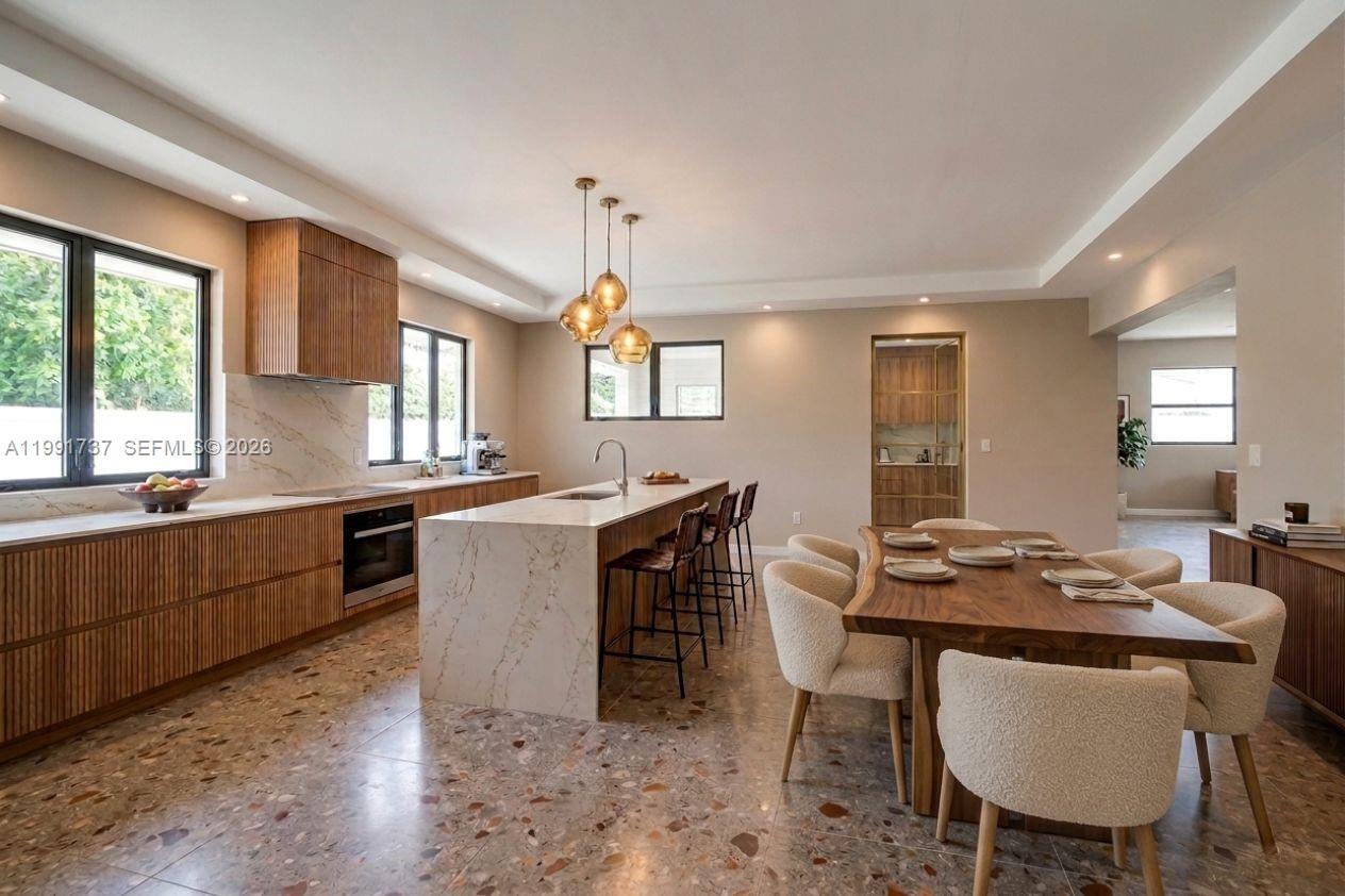 A modern kitchen and dining area features wood cabinetry, a marble island, and terrazzo flooring — 8285 SW 104th St, Miami FL 33156, MIAMI, FL