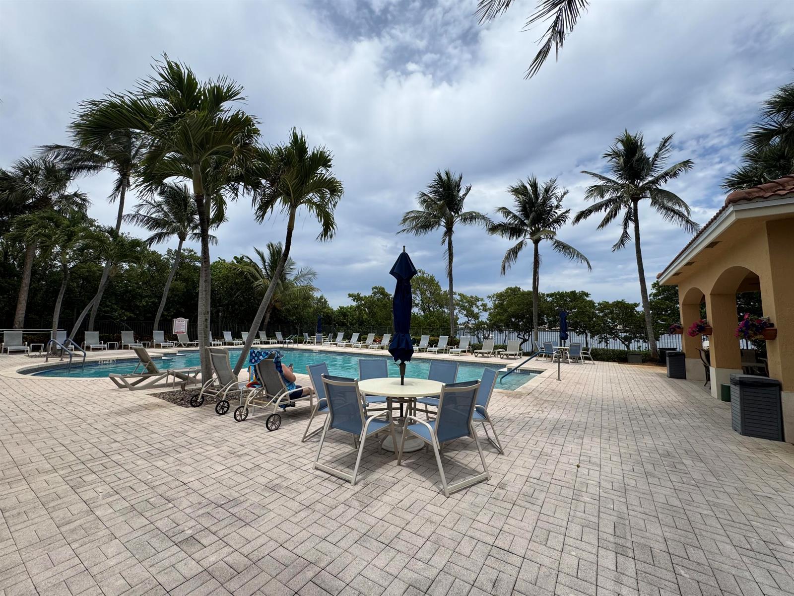 A paved pool deck features a dining set, lounge chairs, and palm trees under a cloudy sky — 700 Scotia Drive # 103, Hypoluxo FL 33462, HYPOLUXO, FL