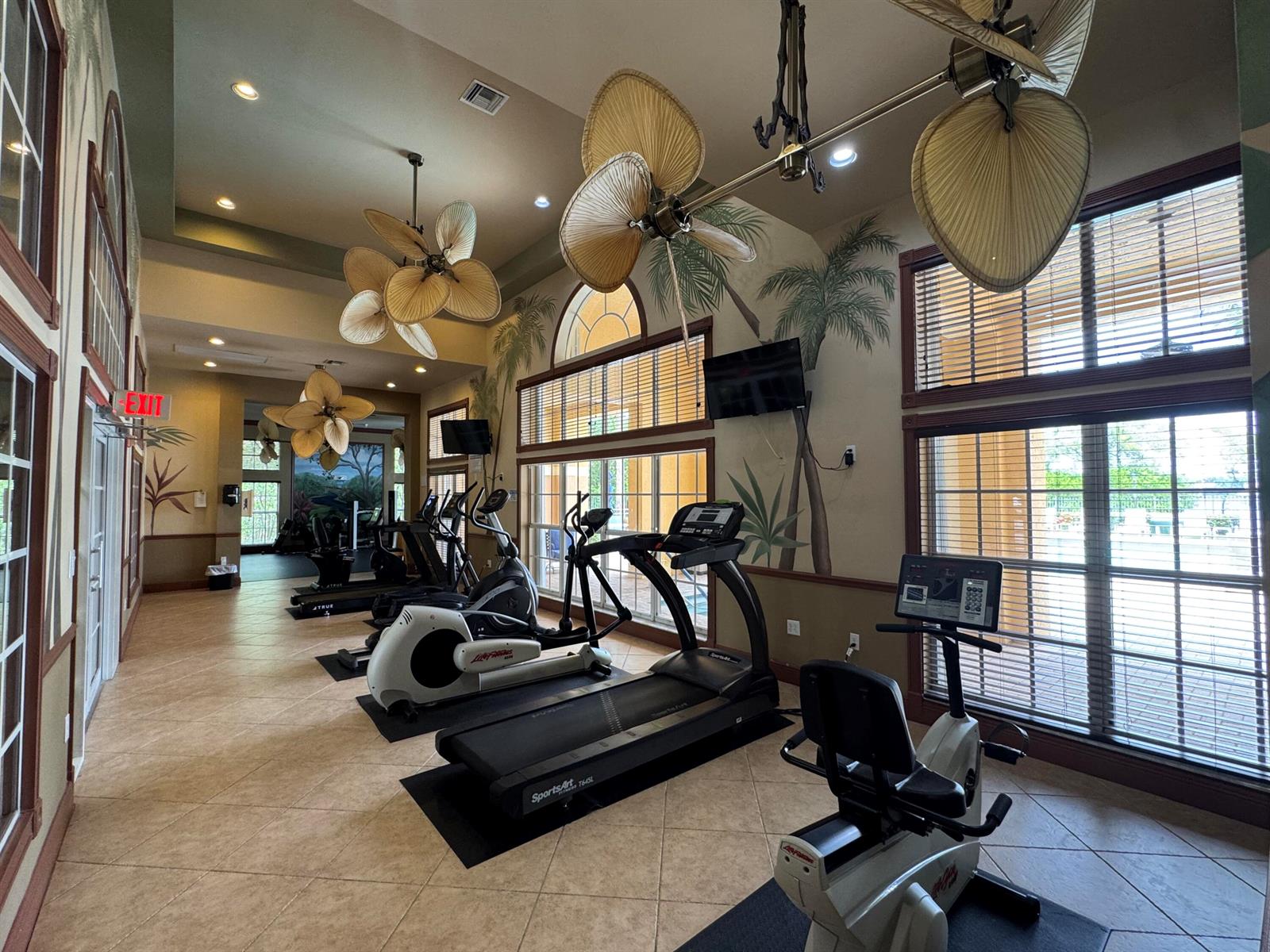 A fitness center features several exercise machines, palm tree wall murals, and unique leaf-shaped fans — 700 Scotia Drive # 103, Hypoluxo FL 33462, HYPOLUXO, FL