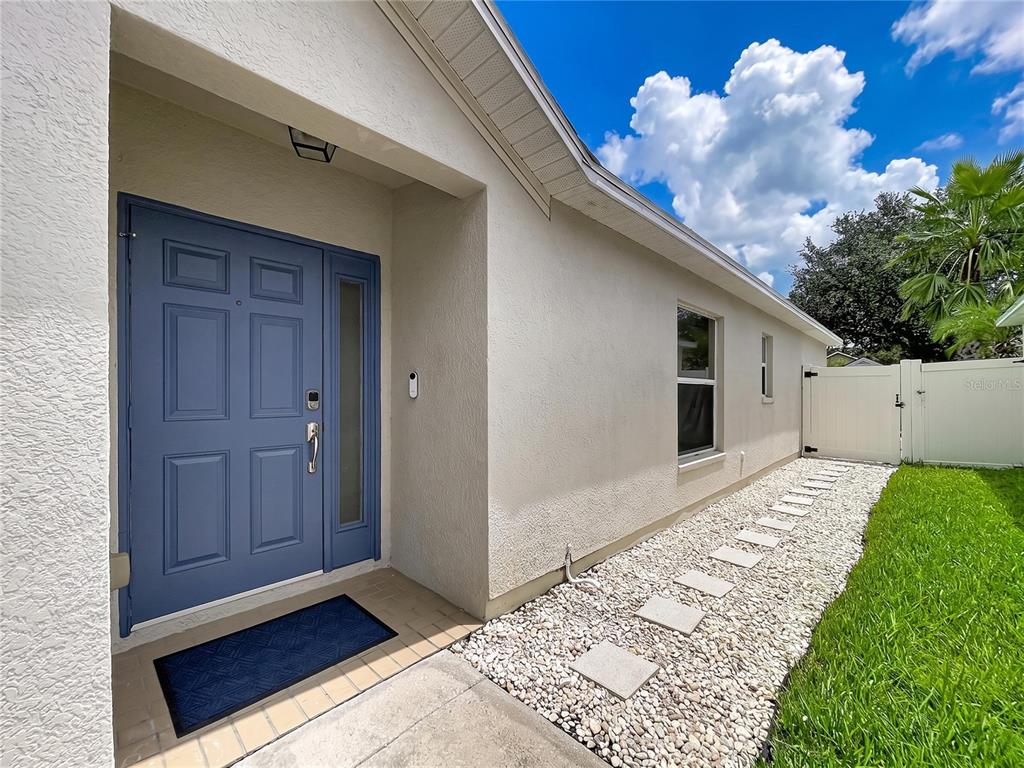 A blue front door with a side light is set into a beige stucco exterior wall — 7710 CARRIAGE POINTE DRIVE, GIBSONTON, FL