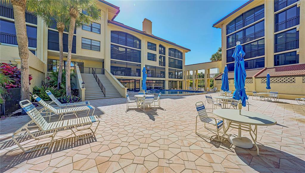 A paved pool deck features lounge chairs, tables with blue umbrellas, and a swimming pool — 8710 MIDNIGHT PASS ROAD UNIT 405, SARASOTA, FL