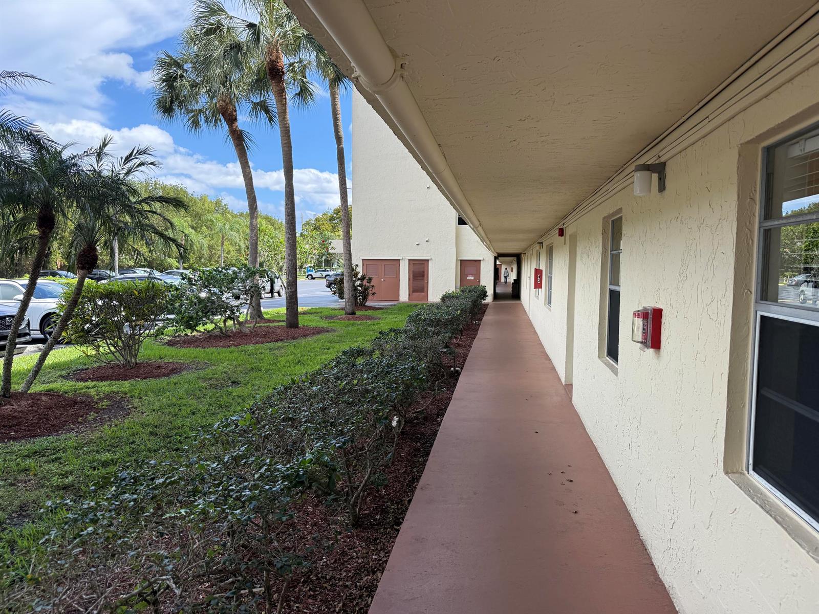 An exterior walkway runs along the side of a beige building with palm trees nearby — 111 SE 3rd Avenue # 108, Dania Beach FL 33004, DANIA BEACH, FL