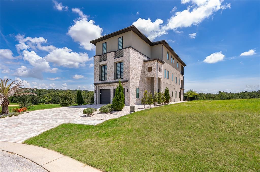 A three-story modern house with stone veneer siding and a paved driveway under a blue sky — 16338 RAVENNA COURT, BELLA COLLINA, FL
