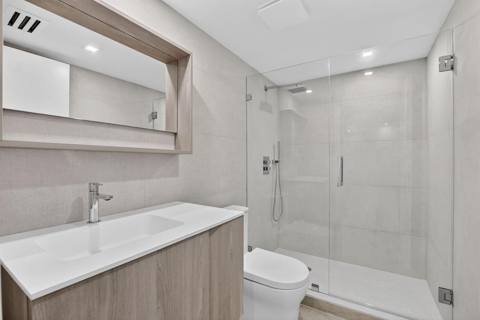 A modern bathroom features a wood vanity, white countertop, and a glass-enclosed walk-in shower — 7835 Lakeside Boulevard # 946, Boca Raton FL 33434, BOCA RATON, FL