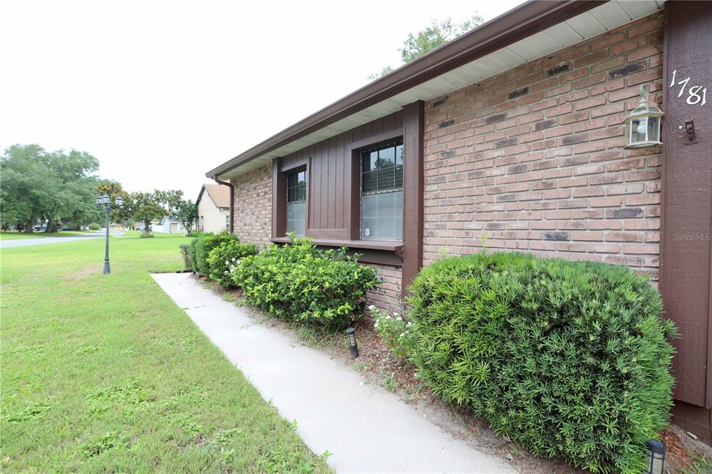 A brick house exterior features a concrete walkway, green bushes, and two windows — 1781 KING CHARLES DRIVE, KISSIMMEE, FL