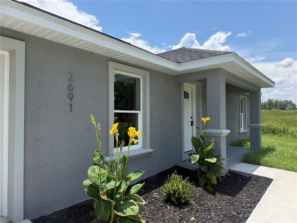 A gray stucco house exterior features a front porch, a window, and yellow flowering plants — 2691 SW 148TH COURT, OCALA, FL