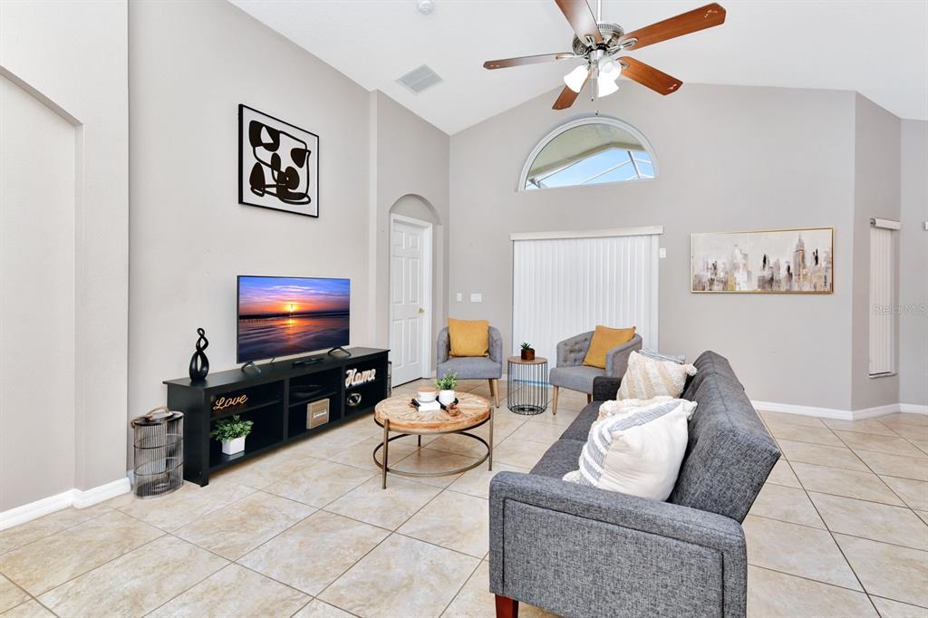 A living room features a vaulted ceiling, tile flooring, a gray sofa, and a television — 4661 CUMBRIAN LAKES DRIVE, KISSIMMEE, FL