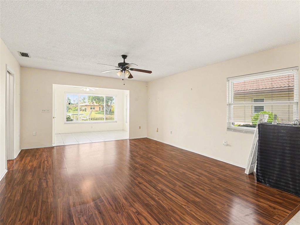 An empty living room features dark wood-look flooring, a ceiling fan, and a sunroom beyond — 6608 AUBURN AVENUE, BRADENTON, FL