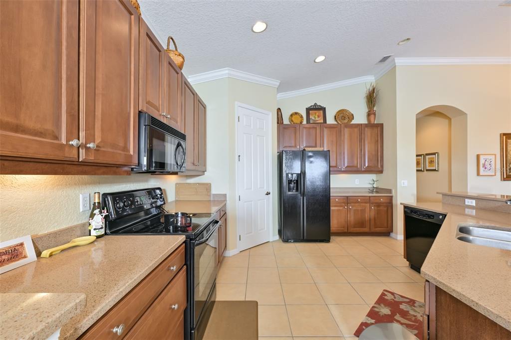 A kitchen features wood cabinets, black appliances, tan countertops, and light-colored tile flooring — 1330 EMERALD DUNES DRIVE, SUN CITY CENTER, FL