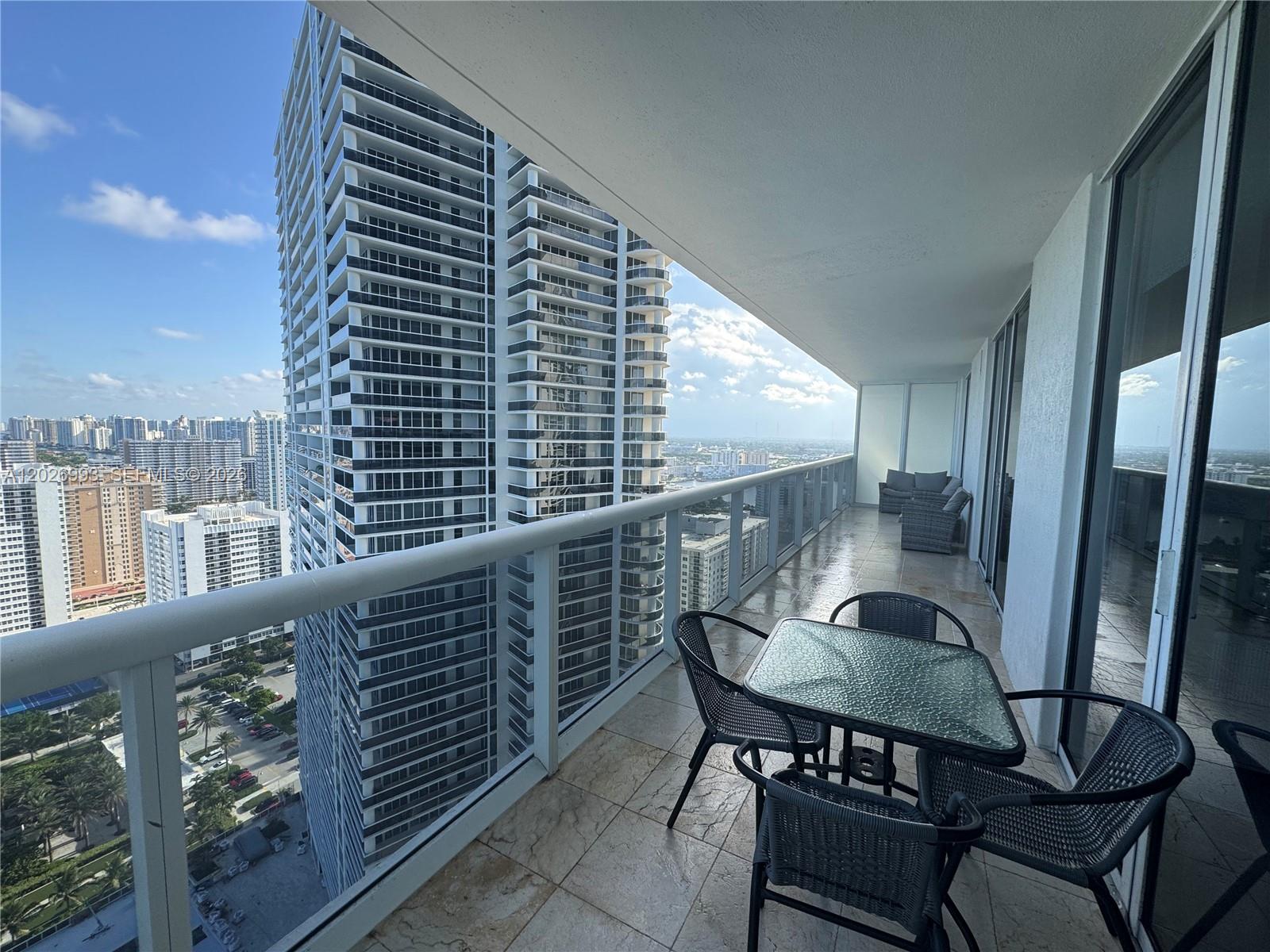A high-rise balcony features a glass-topped table, chairs, and views of surrounding city skyscrapers — 1830 S Ocean Dr # 3003, Hallandale Beach FL 33009, HALLANDALE BEACH, FL