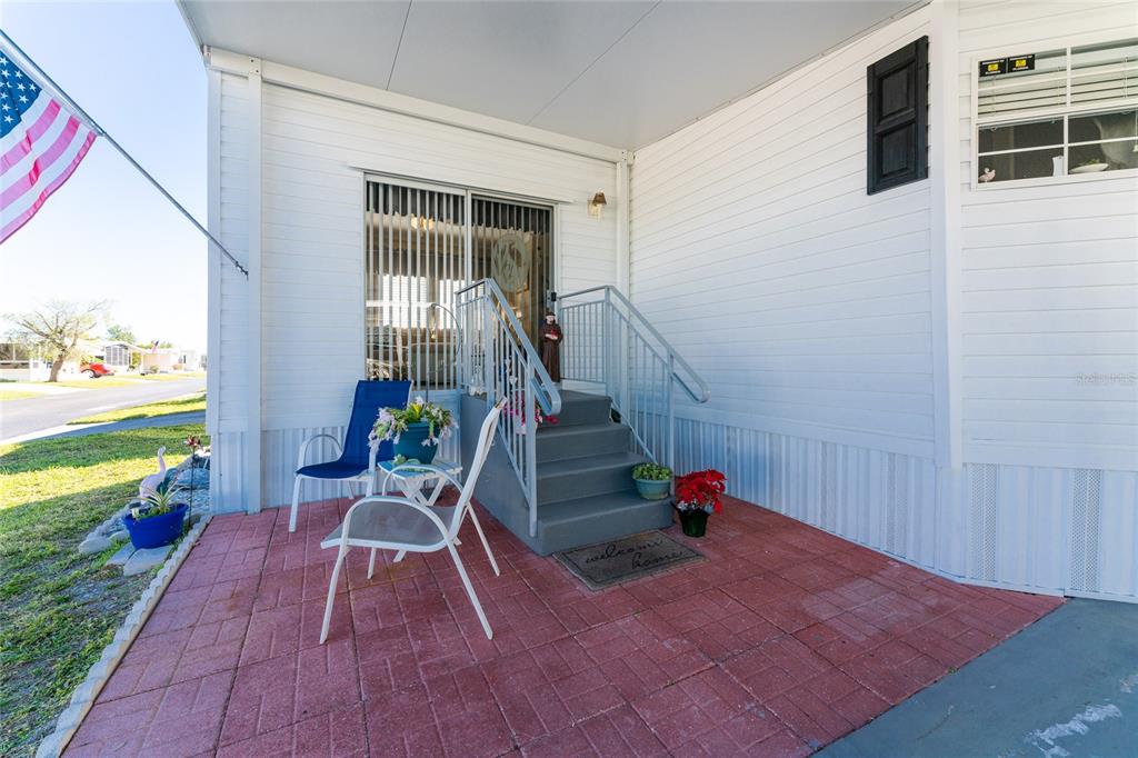 A white mobile home exterior features a red brick patio, stairs with railings, and chairs — 24437 HARBORVIEW ROAD UNIT 62, PUNTA GORDA, FL
